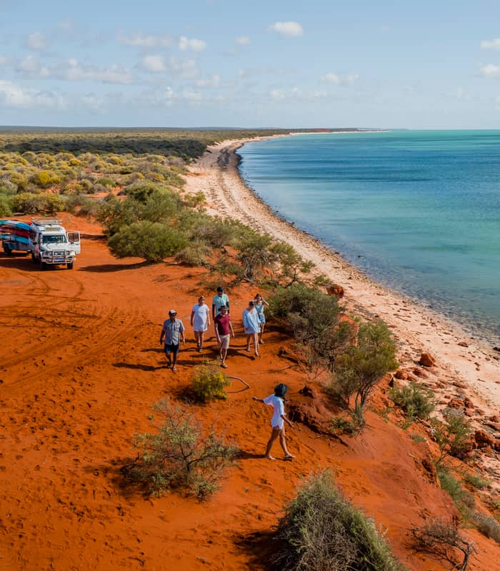 A group of tourists walking along a high red sand cliff next to bright blue sea water.