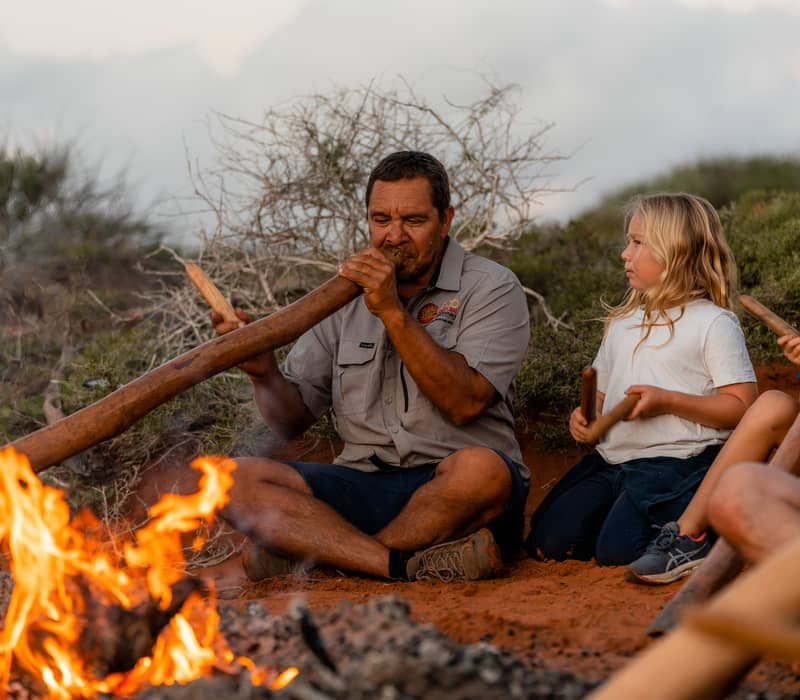 An Indigenous man playing a didgeridoo by a glowing campfire with children sitting nearby.