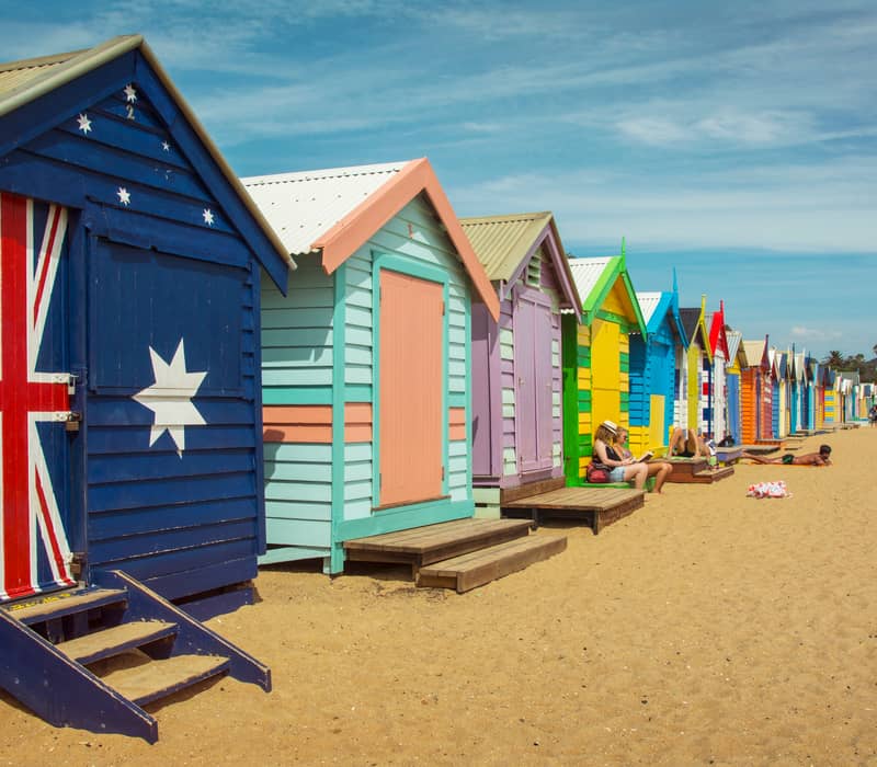 A row of brightly painted wooden beach huts on the sand under a blue sky in Melbourne.