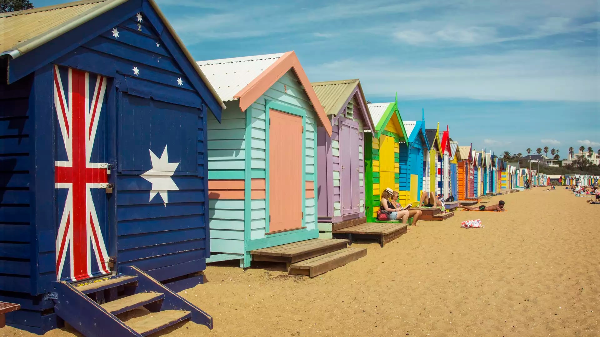 A row of brightly painted wooden beach huts on the sand under a blue sky in Melbourne.