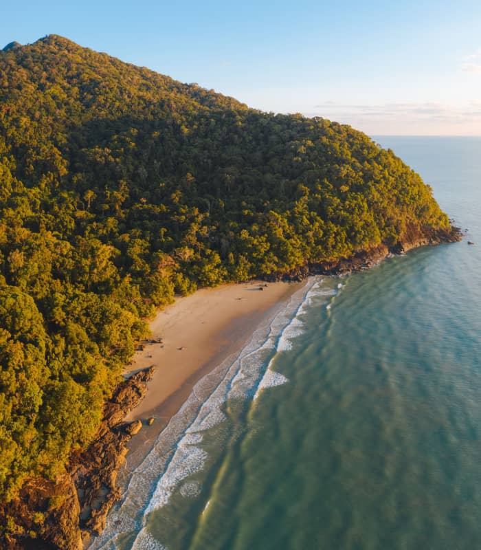 A high-angle aerial view captures a rocky, tree-covered headland at the end of Noah Beach, with the sun casting a warm glow over the dense Daintree Rainforest and the vast ocean.