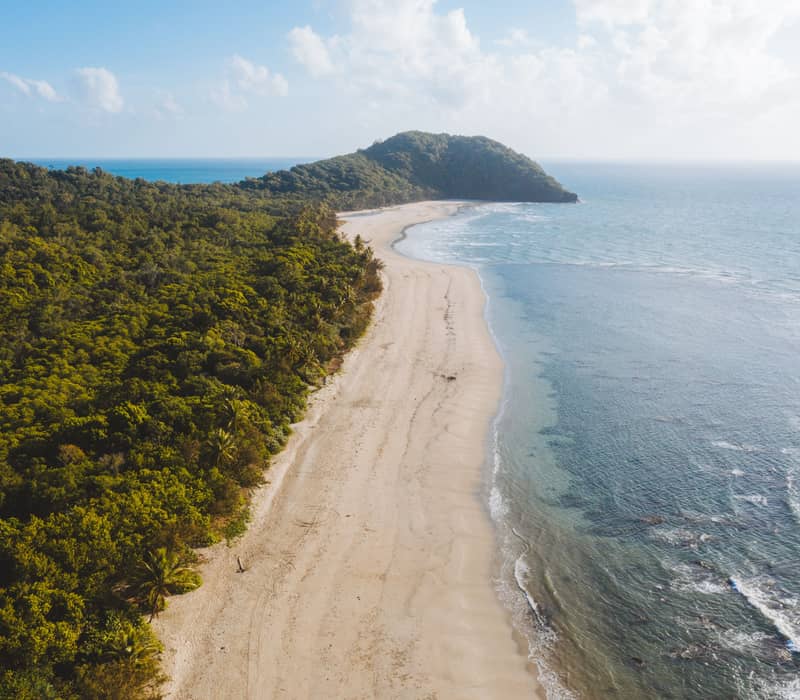 An aerial perspective looking down the long, curving white sand beach of Myall Beach, bordered by thick green jungle and leading to the distant forested point of Cape Tribulation.