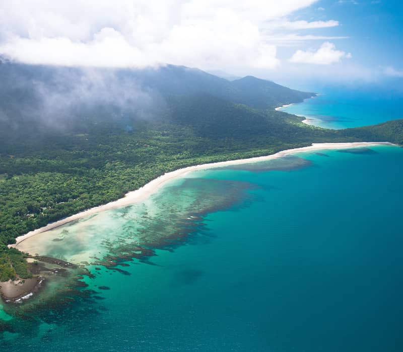 A spectacular wide aerial view shows white clouds and mist clinging to the steep green peaks of the Daintree Rainforest mountains as they meet the vibrant blue waters and reefs of the Coral Sea.