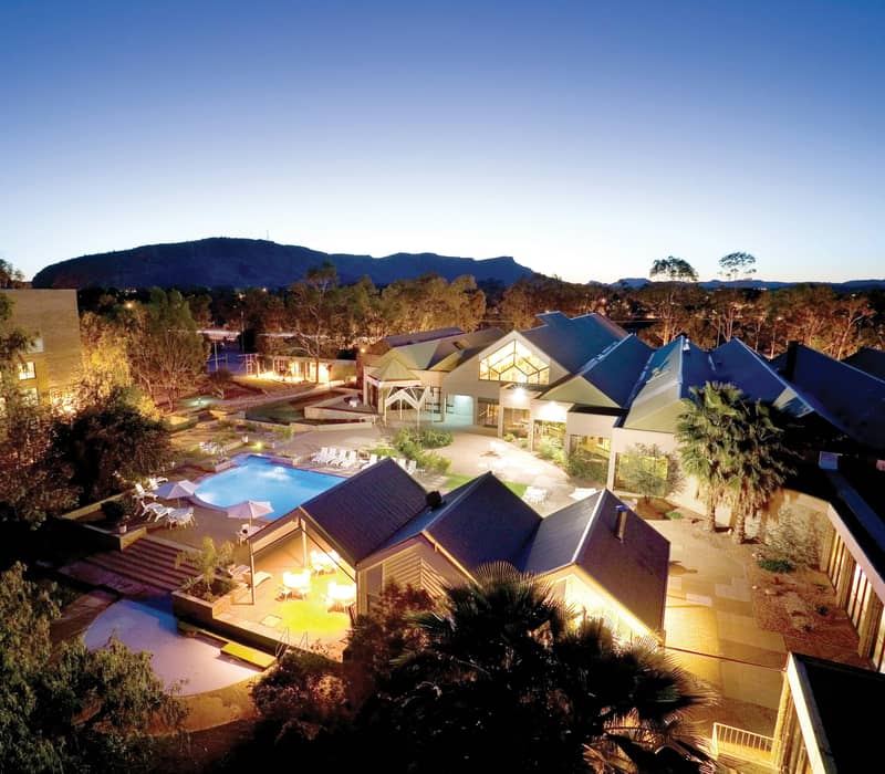 A vibrant peacock with a long tail walks past a large blue swimming pool and lounge chairs under a bright blue sky at a resort in Alice Springs.