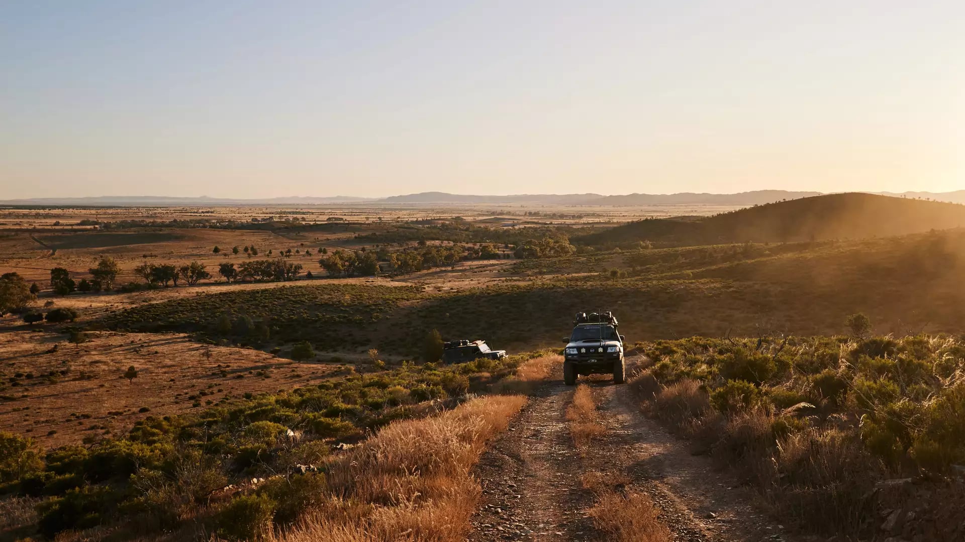 Two 4WD vehicles driving along a dusty mountain ridge track in the Flinders Ranges during sunset.