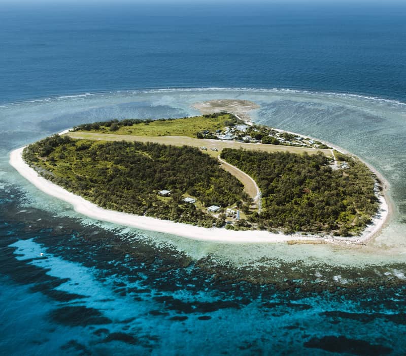 A wide aerial perspective shows the lush green Lady Elliot Island coral cay with its central grass airstrip, surrounded by vibrant turquoise reef and deep blue ocean.