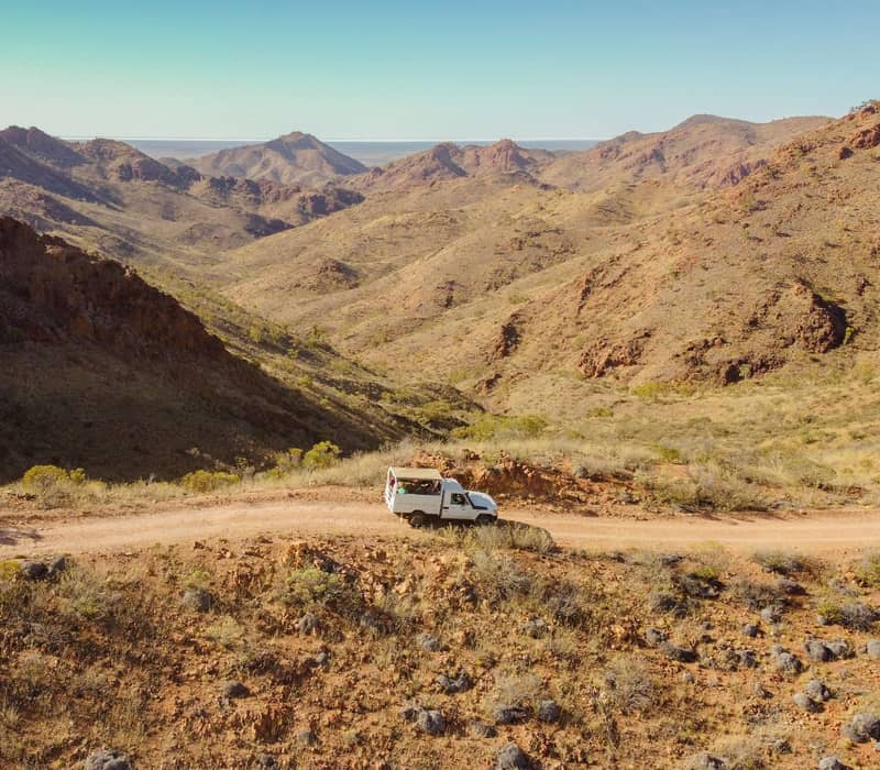 A white safari-style 4WD vehicle parked on a narrow mountain ridge in a desert landscape.