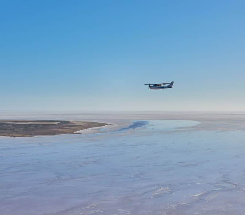 A scenic flight plane soaring over a blue and white crystalline salt lake in the Australian outback.
