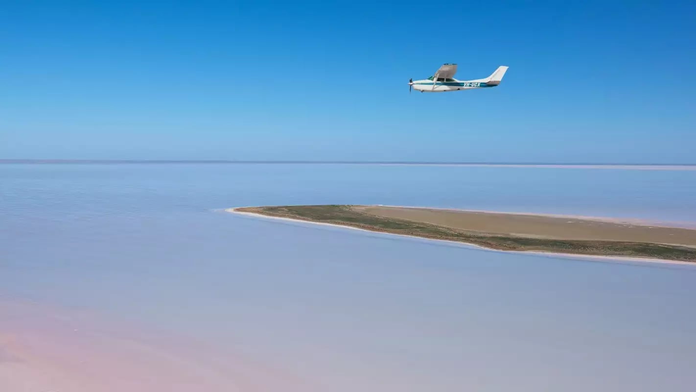 A small white scenic plane flying over the vast pink and white salt crust of Lake Eyre.