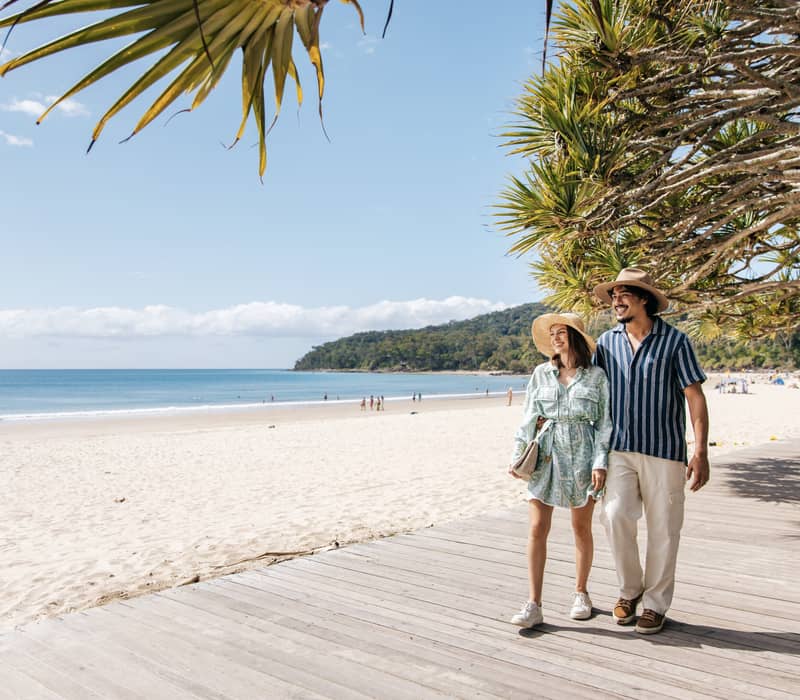 Couple walking along a wooden boardwalk beside a white sandy beach with turquoise water under tropical trees.