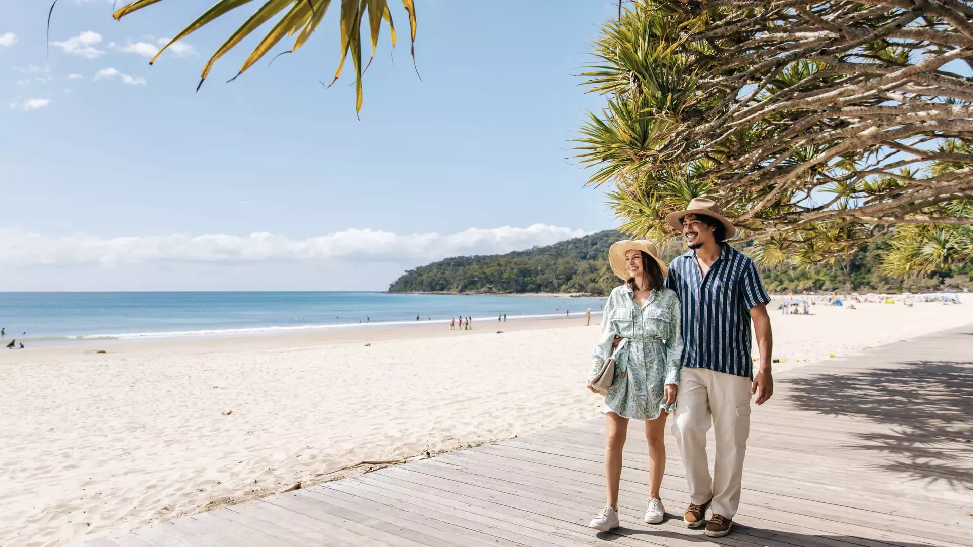 Couple walking along a wooden boardwalk beside a white sandy beach with turquoise water under tropical trees.