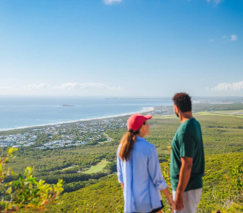 Two hikers standing on a mountain peak looking out over the coastline and green landscape below.