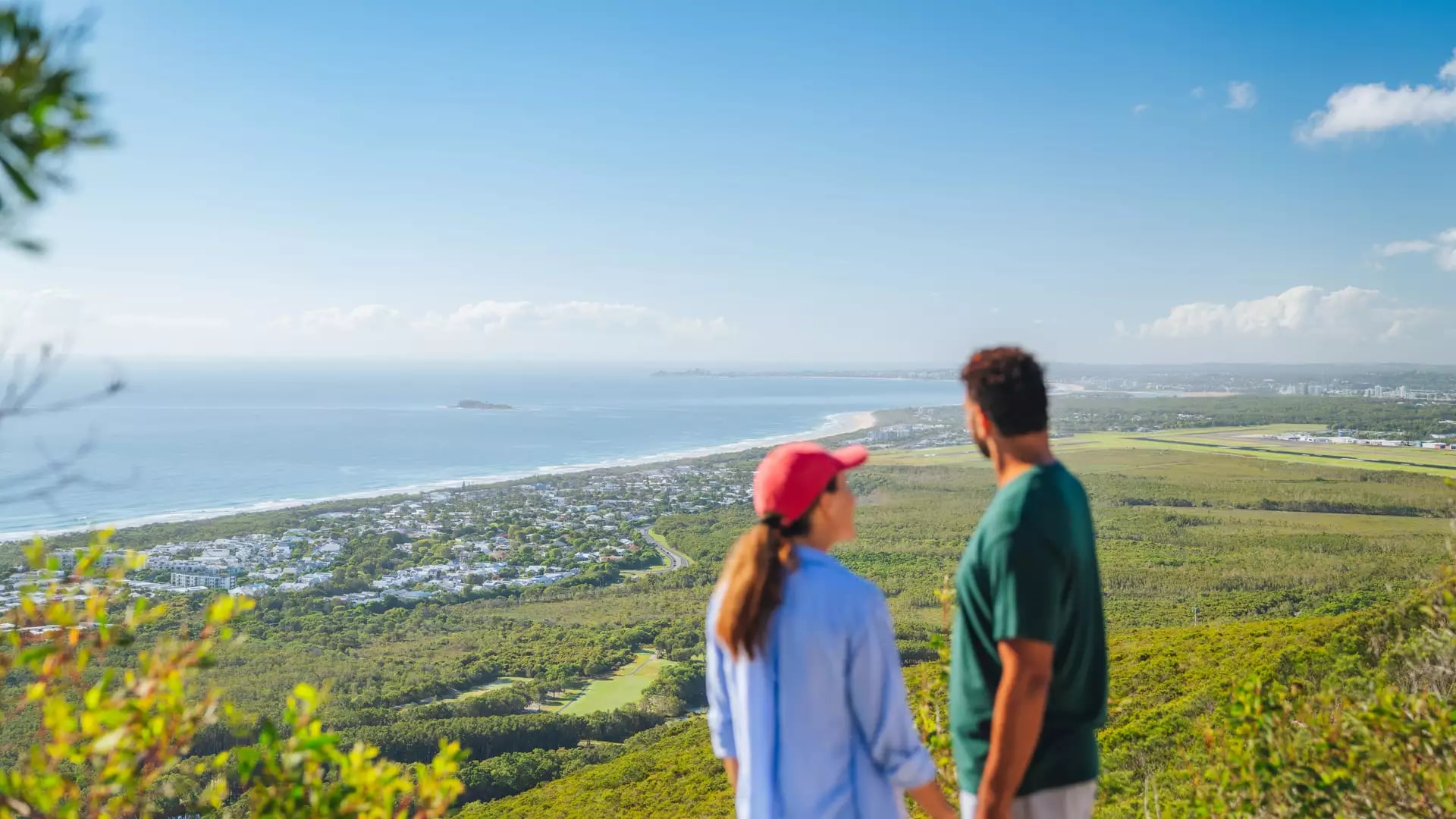 Two hikers standing on a mountain peak looking out over the coastline and green landscape below.