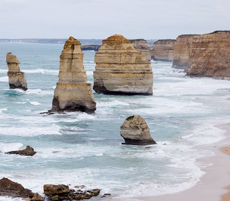 Several large limestone rock towers standing in turquoise water near a sandy beach and high golden cliffs under an overcast sky.