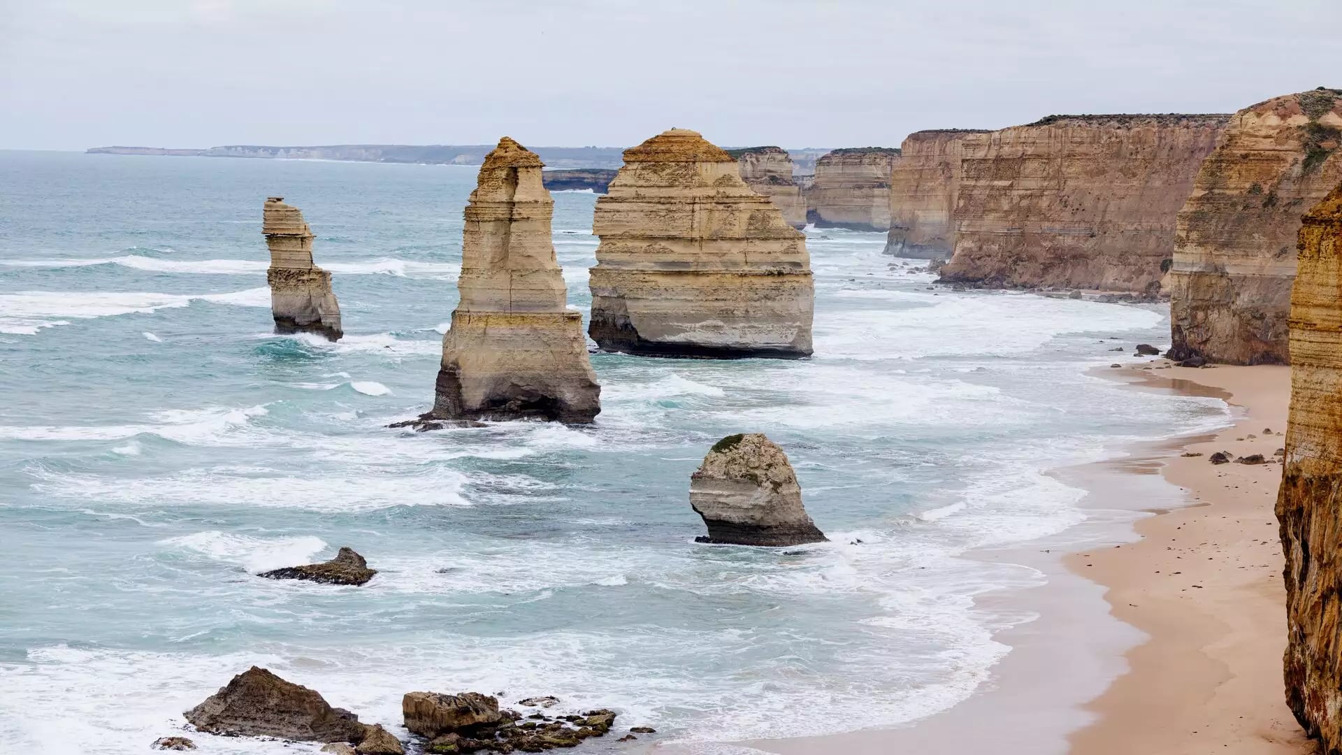 Several large limestone rock towers standing in turquoise water near a sandy beach and high golden cliffs under an overcast sky.