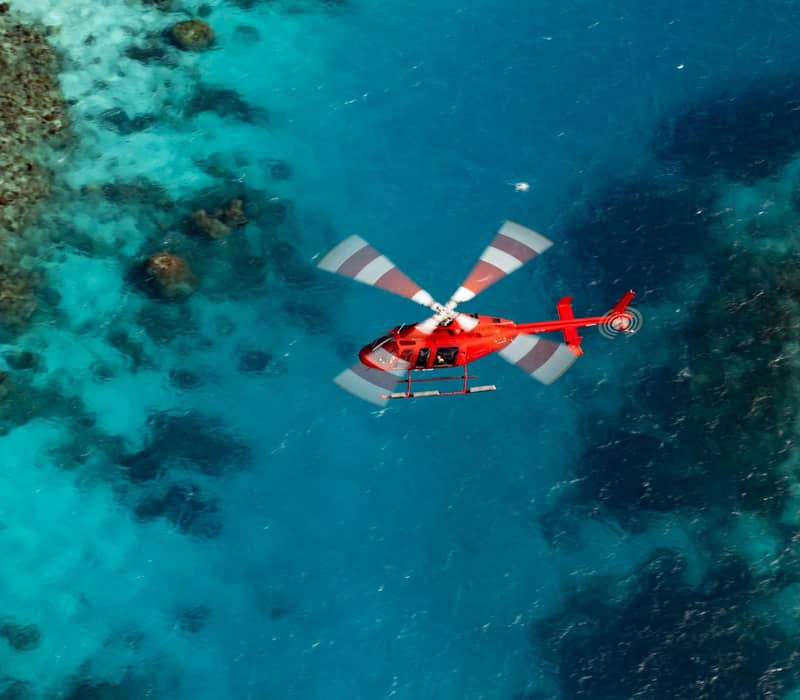 Red helicopter flying over turquoise ocean with visible coral reefs beneath