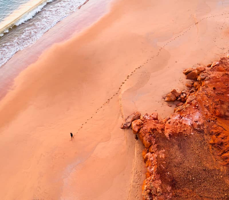 A high-angle shot of a man standing on a rugged, dark red rock ledge overlooking a sandy beach and incredibly clear, light blue water.