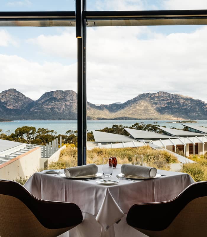 A fine dining table set for two with white linen in front of a large window overlooking the Tasmanian coastline and mountains.