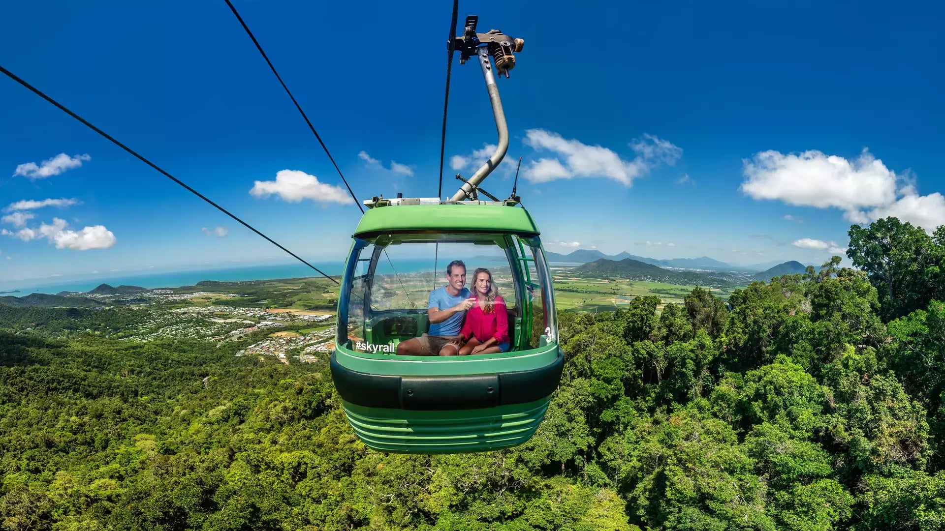 Travelers in a green Skyrail gondola looking out over the Cairns rainforest canopy and ocean.