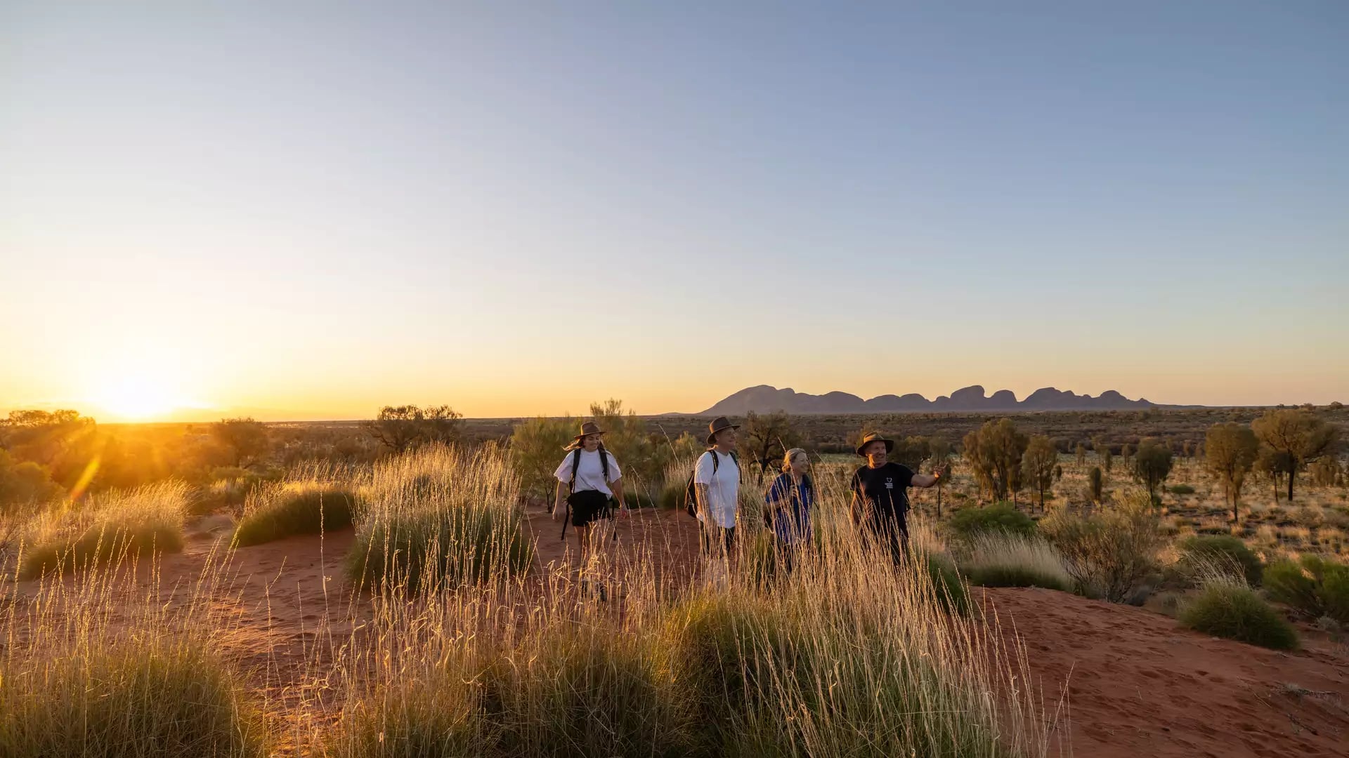Hikers walking through red sand dunes and tall grass during a golden sunrise with the Olgas in the background.