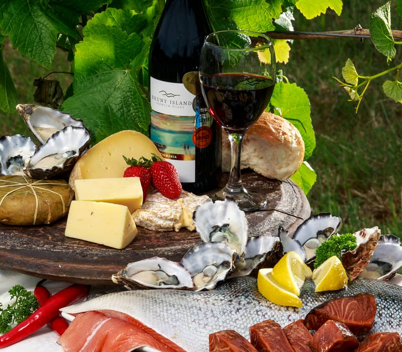 A platter of fresh Tasmanian oysters, smoked salmon, local cheeses, and red wine against a vineyard background.