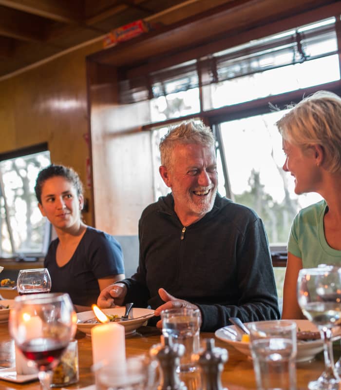 Hikers laughing and sharing a meal with red wine at a wooden table inside a rustic lodge.