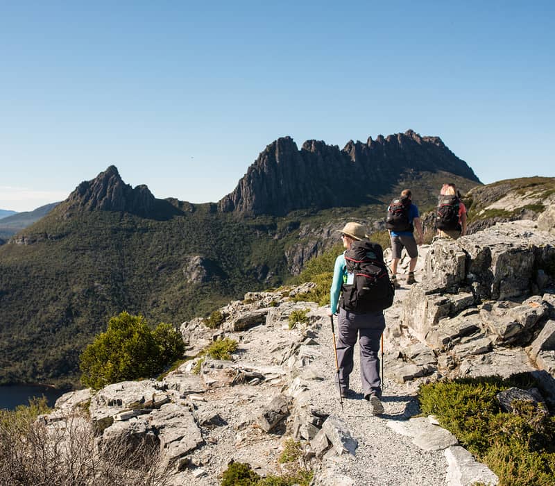 Hikers walking along a narrow rocky path overlooking a deep valley and distant blue mountain ranges.
