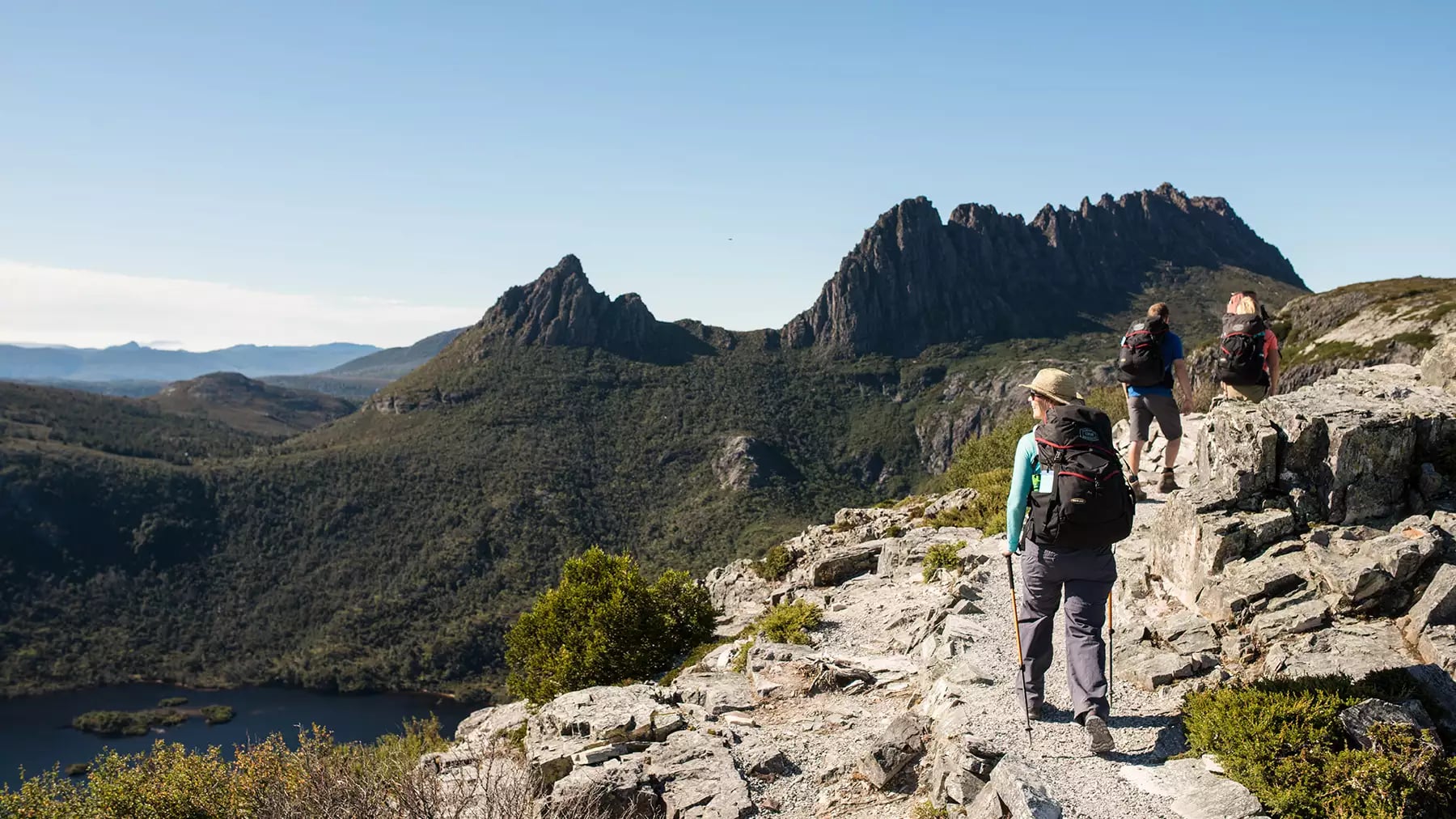 Hikers walking along a narrow rocky path overlooking a deep valley and distant blue mountain ranges.