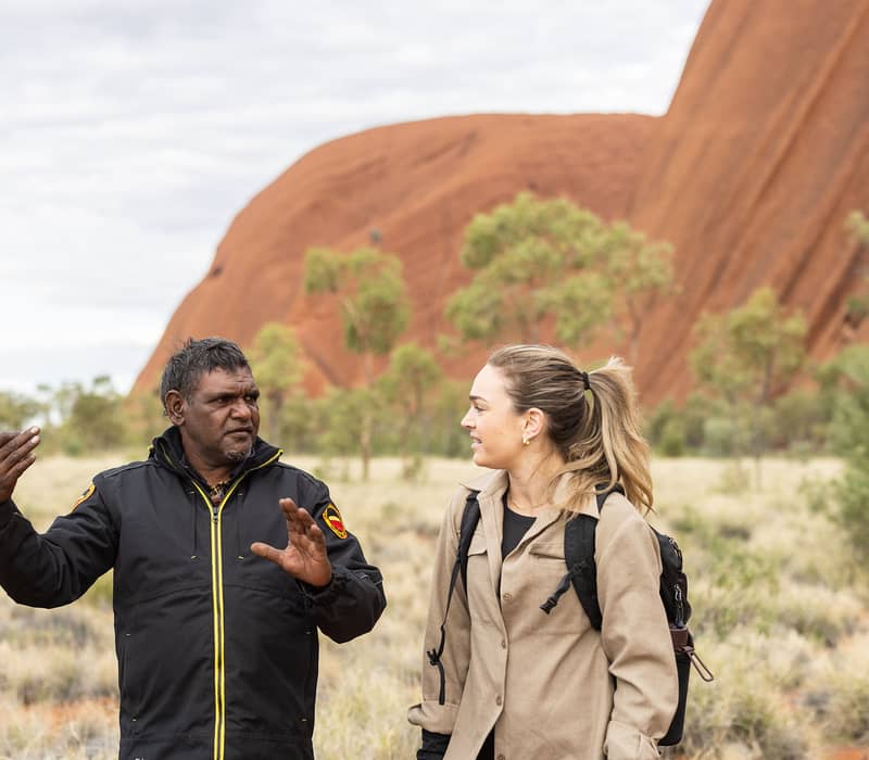 An Aboriginal guide talking to a female hiker in front of the massive red rock walls of Uluru.