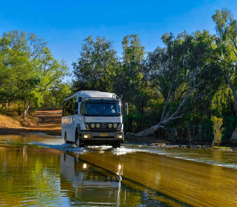 A white Outback Spirit 4WD bus crosses a shallow, sunlit river on a dirt track