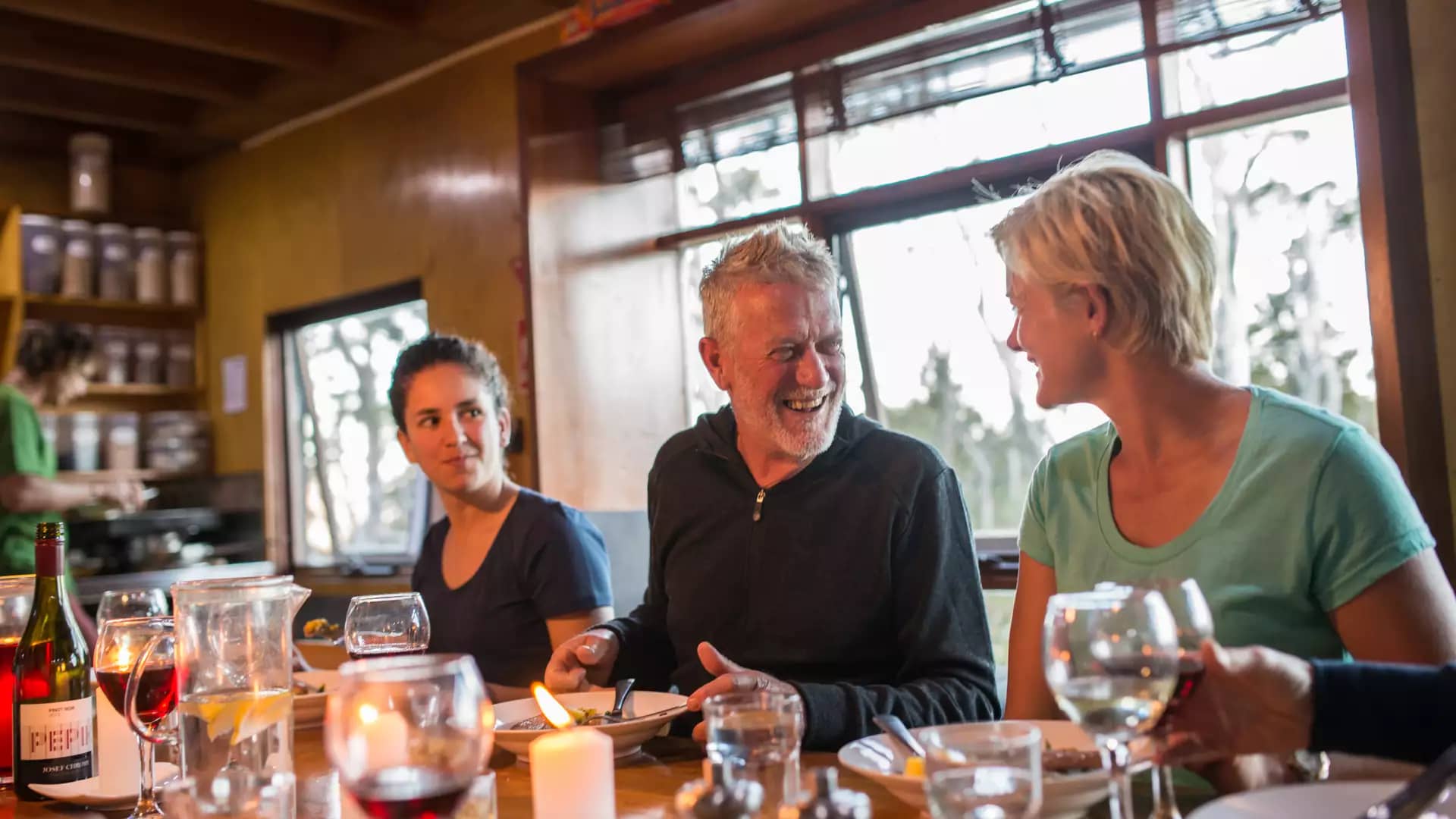 Hikers laughing and sharing a meal with red wine at a wooden table inside a rustic lodge.