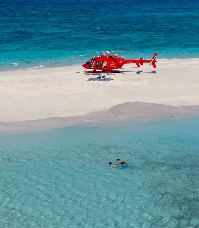 A red helicopter landed on a small white sand cay in the middle of a turquoise ocean with people snorkeling nearby.
