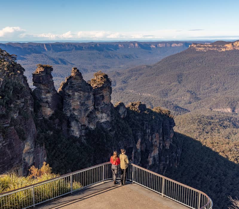 Two people standing at a lookout railing facing the Three Sisters rock formations and vast blue-toned valley.