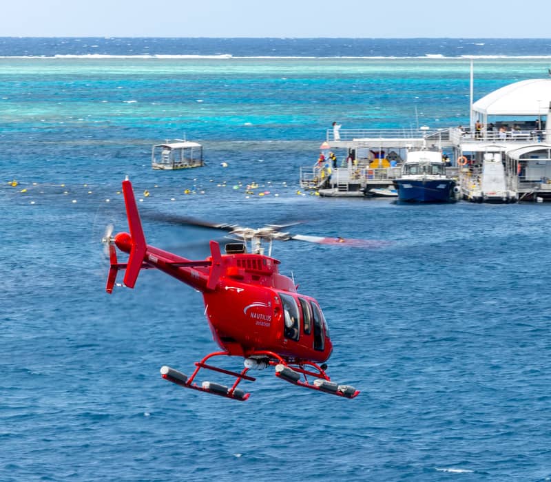 A red helicopter flying over the blue waters of the Great Barrier Reef toward a tourist pontoon and boat.