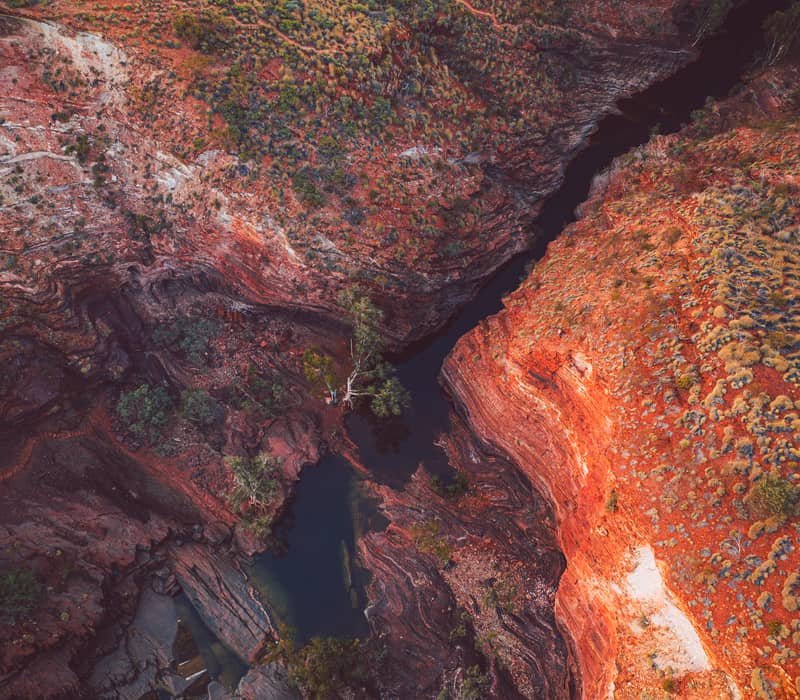 An aerial photograph looking directly down into a narrow, dark blue river winding through a vast landscape of jagged, iron-red rock formations and sparse green vegetation.