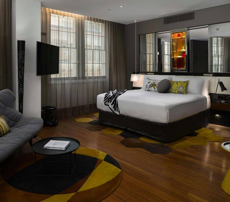 Modern hotel room with king bed, gray sofa, round rug, and large windows with sheer curtains.