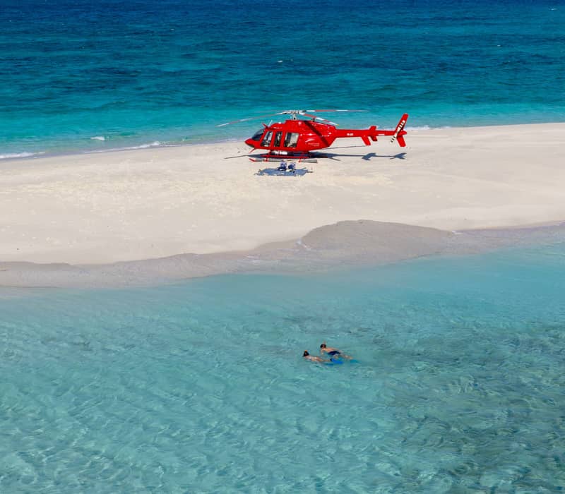 A red helicopter landed on a small white sand cay in the middle of a turquoise ocean with people snorkeling nearby.
