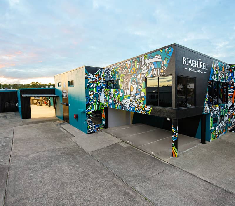 A modern distillery building with colorful abstract murals on the exterior walls under a sunset sky in Caloundra.