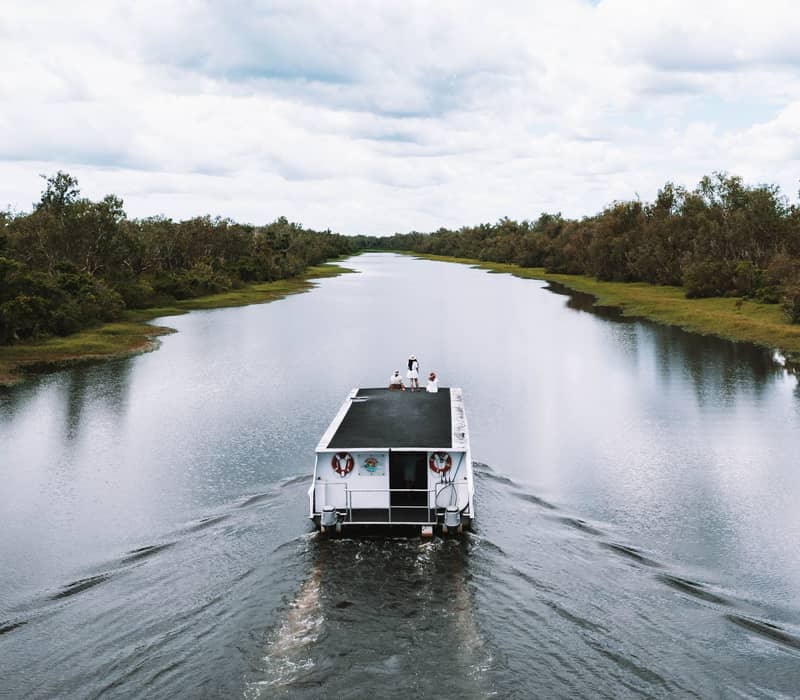A high-altitude drone view shows a white boat cruising down the center of a wide, calm river flanked by dense green bushland under a cloudy sky in the Top End.
