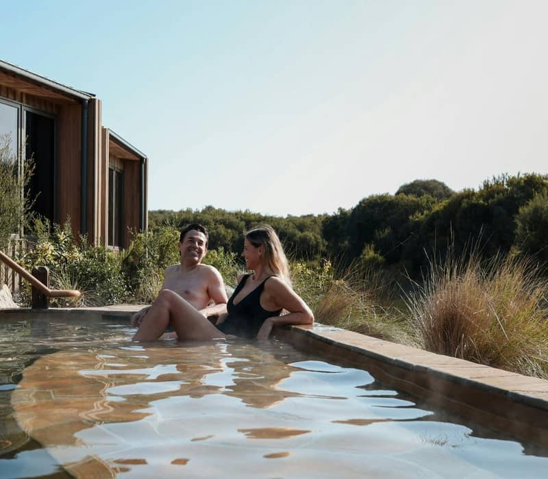 Couple sitting in outdoor hot springs pool with natural greenery and lodge building nearby.