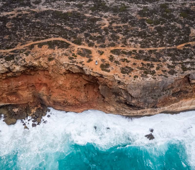 A white 4WD vehicle sits on the rugged, orange clifftops of Toolina Cove overlooking the turquoise Southern Ocean and white crashing waves in Esperance, Western Australia.