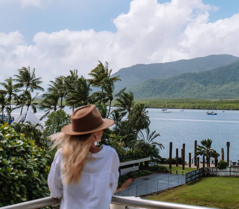 A woman in a sun hat overlooks the blue waters of Cairns harbor and the surrounding green mountains.
