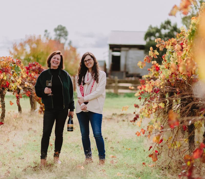 Two women holding a bottle and glass of red wine smiling in a vineyard with bright autumn leaves.