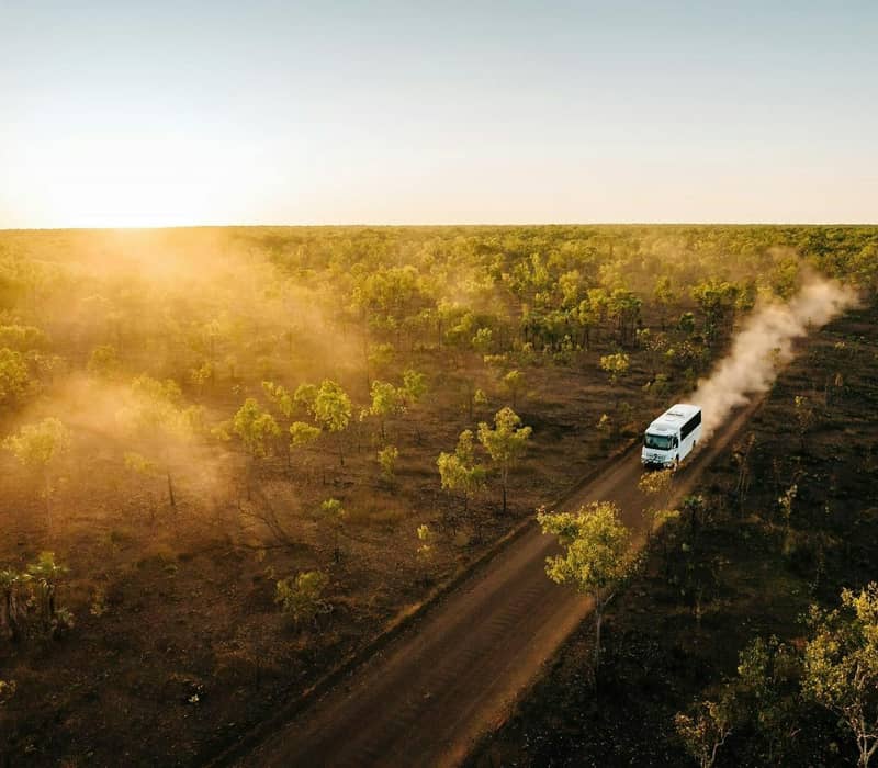 An aerial view of an Outback Spirit 4WD safari coach traveling along a remote dirt track during a Kimberley sunset. The golden light illuminates the dust trail and the vast, wooded savanna landscape.