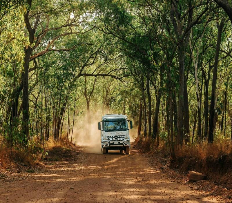 An Outback Spirit safari coach navigates a narrow dirt road framed by a dense canopy of eucalyptus and tropical trees. This perspective highlights the immersive nature of 4WD touring through the remote regions of Western Australia.
