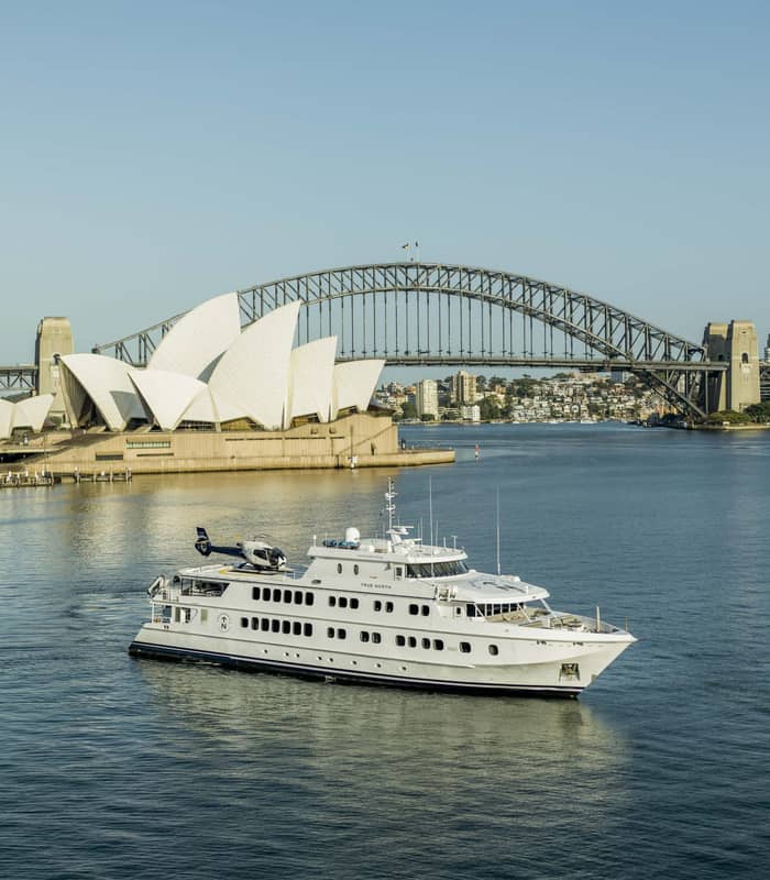 A wide landscape shot of the white True North on calm blue water, framed by the white sails of the Opera House and the steel arch of the Harbour Bridge.