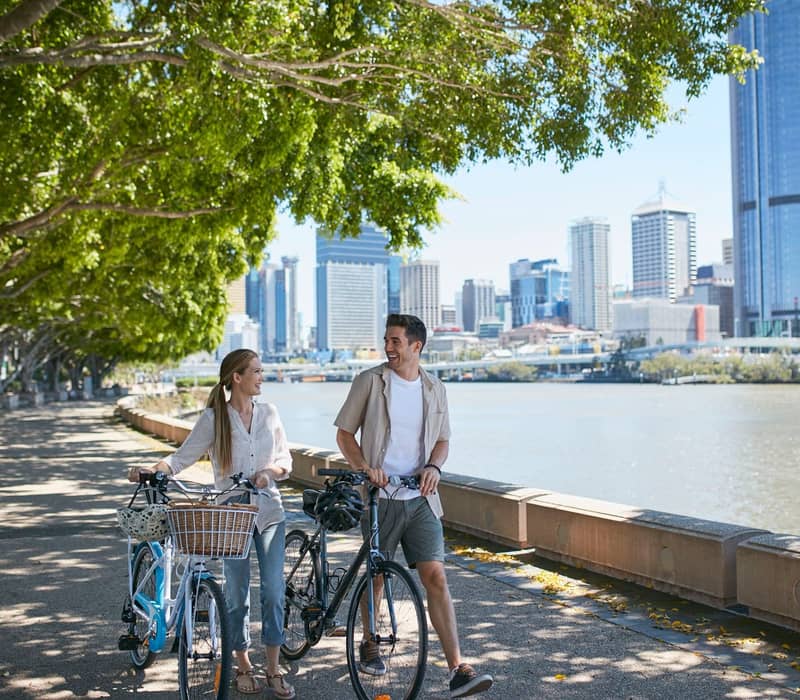 Man and woman walking with bicycles along a tree-lined riverside path with Brisbane city buildings in the background.