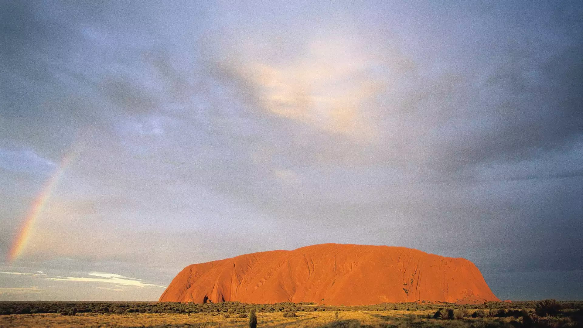 The red monolith of Uluru in the distance with a bright rainbow appearing on the left side under a cloudy sky.