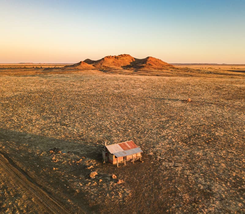 Aerial view of a small rusted iron hut in a vast dry field leading to a flat-topped mountain at sunset in Queensland.