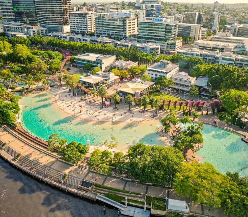 Drone shot of the blue man-made lagoon at South Bank Parklands surrounded by sand and city buildings.